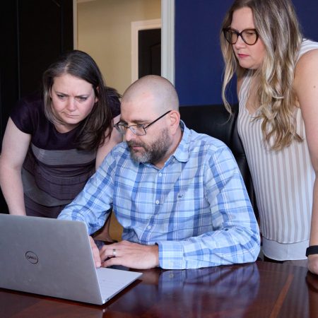 Cynthia, Geoff, and Tayla gathered around a laptop in an office, focused on the screen while discussing work.