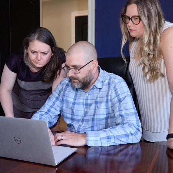 marketing-analytics-and-reporting-with-cut-throat Cynthia, Geoff, and Tayla gathered around a laptop in an office, focused on the screen while discussing work.