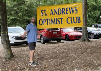 Man posing outdoors in front of a large yellow banner that reads “St. Andrews Optimist Club,” with parked cars in the background.